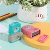 A set of pink and teal custom kids name stamps resting on a yellow surface next to a white potted plant.
