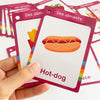 A hand holds a pink flash card showing a hot dog, part of a set of bilingual vocabulary cards.