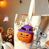 A beige tote bag features embroidered Halloween plush charms including a purple ghost, an orange pumpkin, and a white bat ske