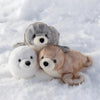 A grouping of three plush toys: a gray seal, a white seal, and a light brown seal, lying in the snow.