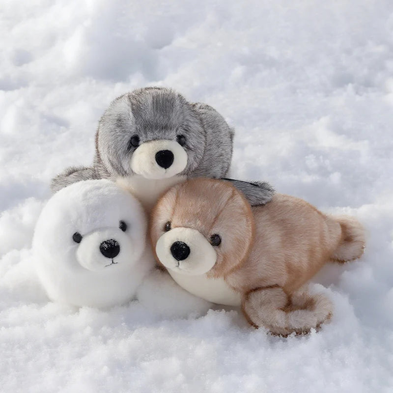 A grouping of three plush toys: a gray seal, a white seal, and a light brown seal, lying in the snow.