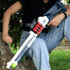 A person holds a white and black soft bullet shotgun toy with an ejecting shell action against a tree trunk.