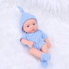 A realistic soft vinyl reborn baby doll wearing a light blue knitted hat and outfit, lying on white fluffy material.