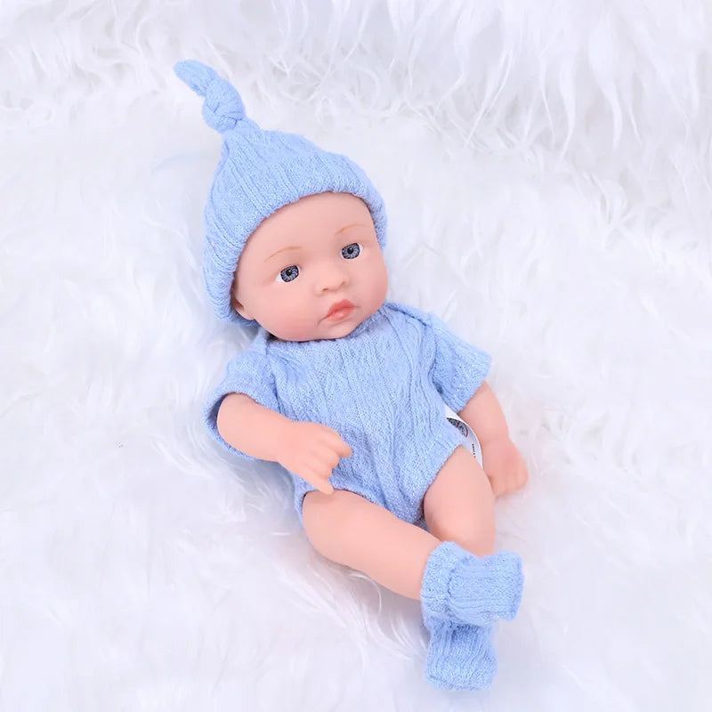 A realistic soft vinyl reborn baby doll wearing a light blue knitted hat and outfit, lying on white fluffy material.