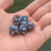 A hand holds a set of seven iridescent acrylic polyhedral dice on a green background.