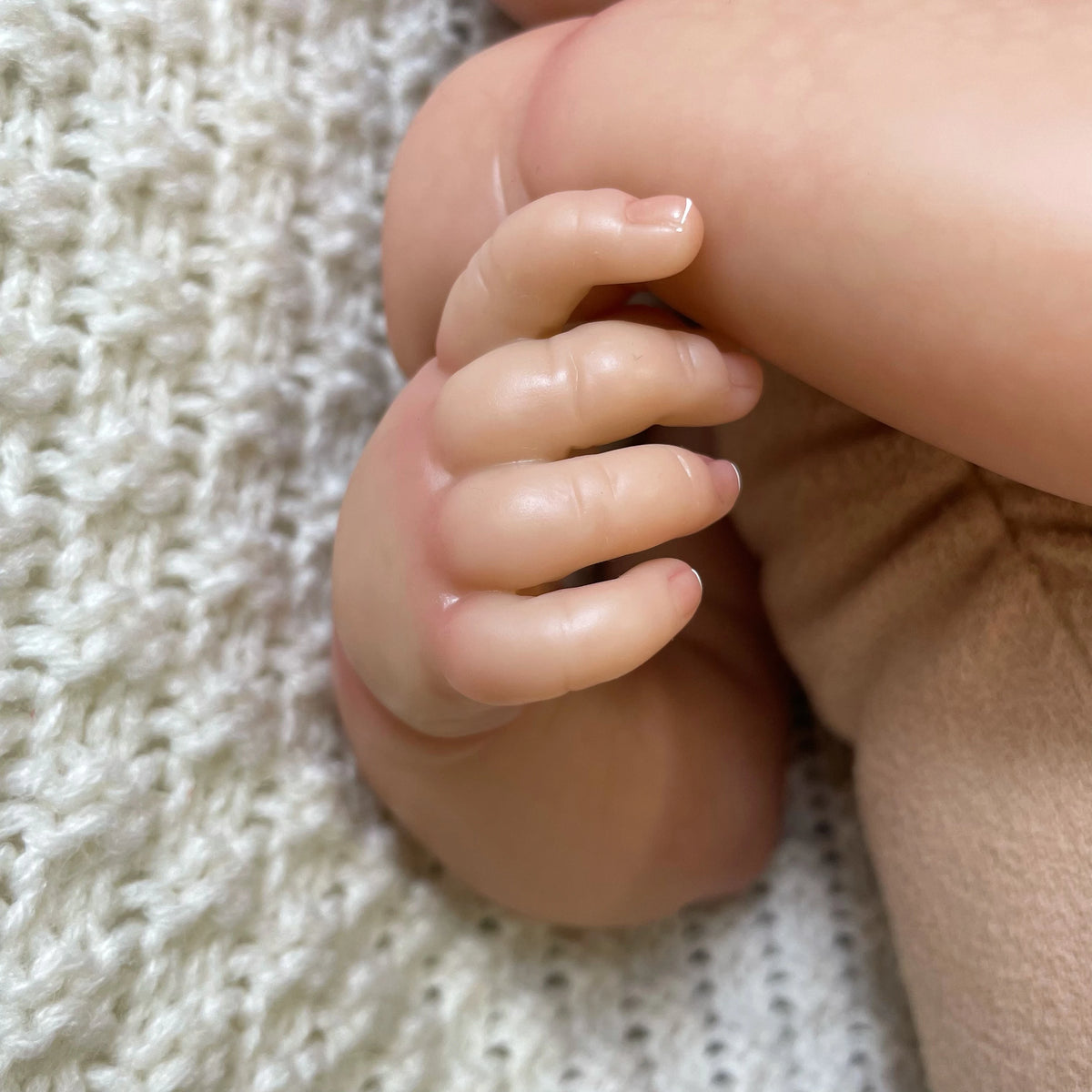A close-up of a realistic reborn baby doll's hand and forearm resting on a knitted fabric surface.