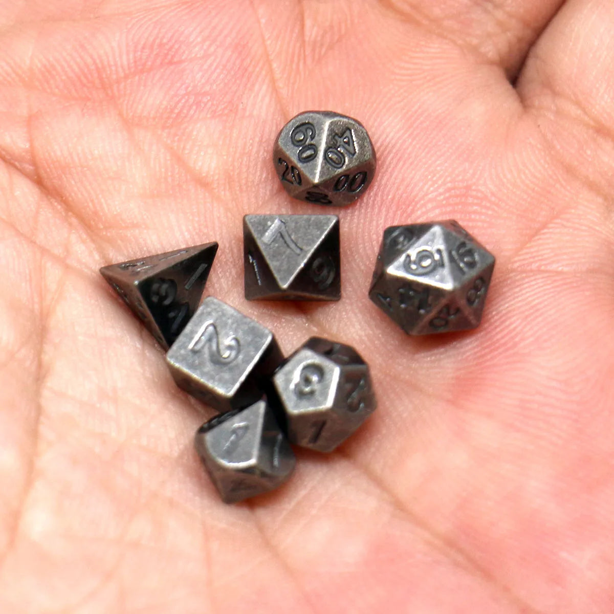 A hand holds a set of seven antique metal polyhedral dice, including a d20 and d10.