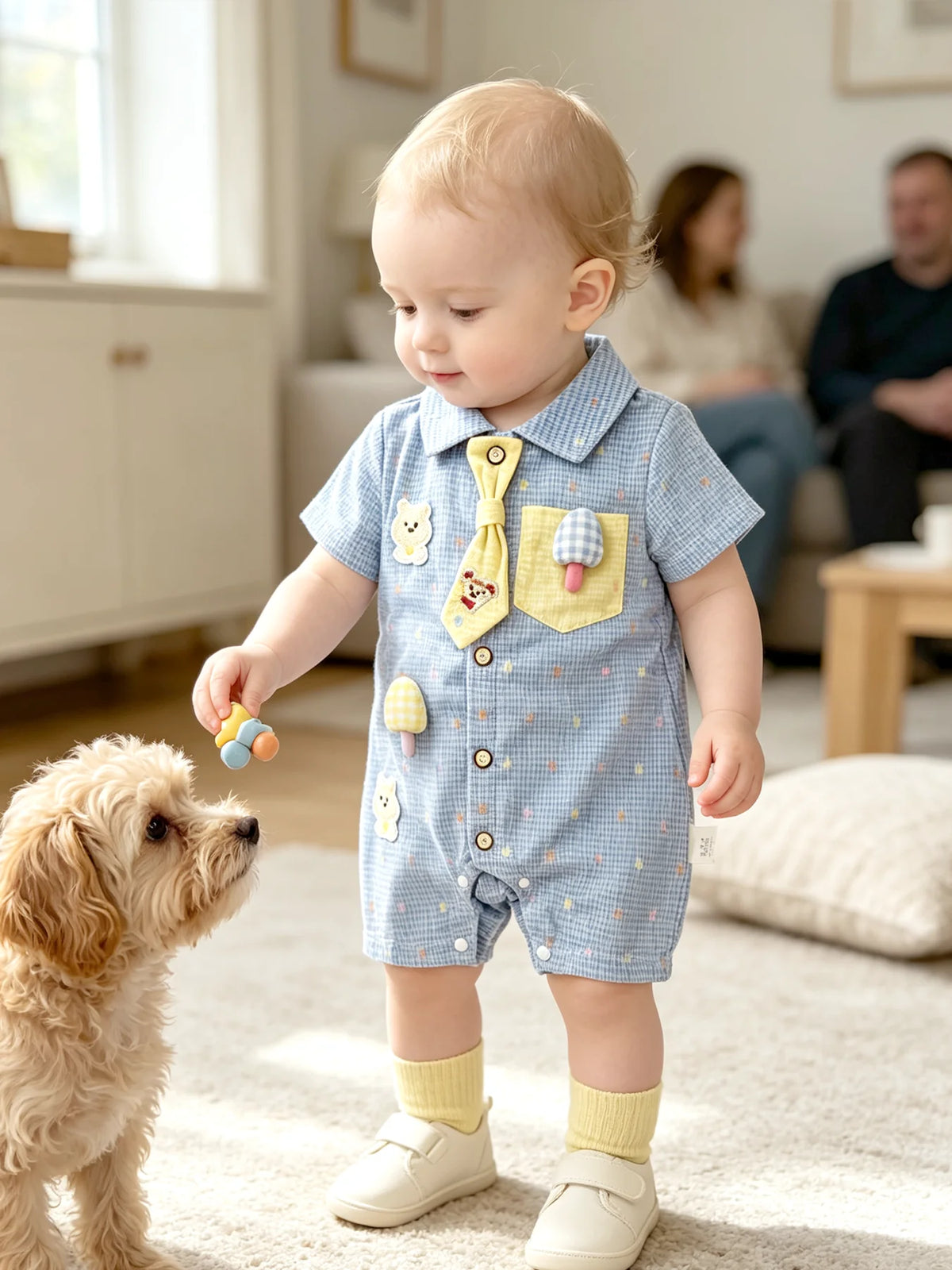 A baby boy wearing a blue patterned short-sleeve romper with bear and mushroom appliqués standing on a rug.