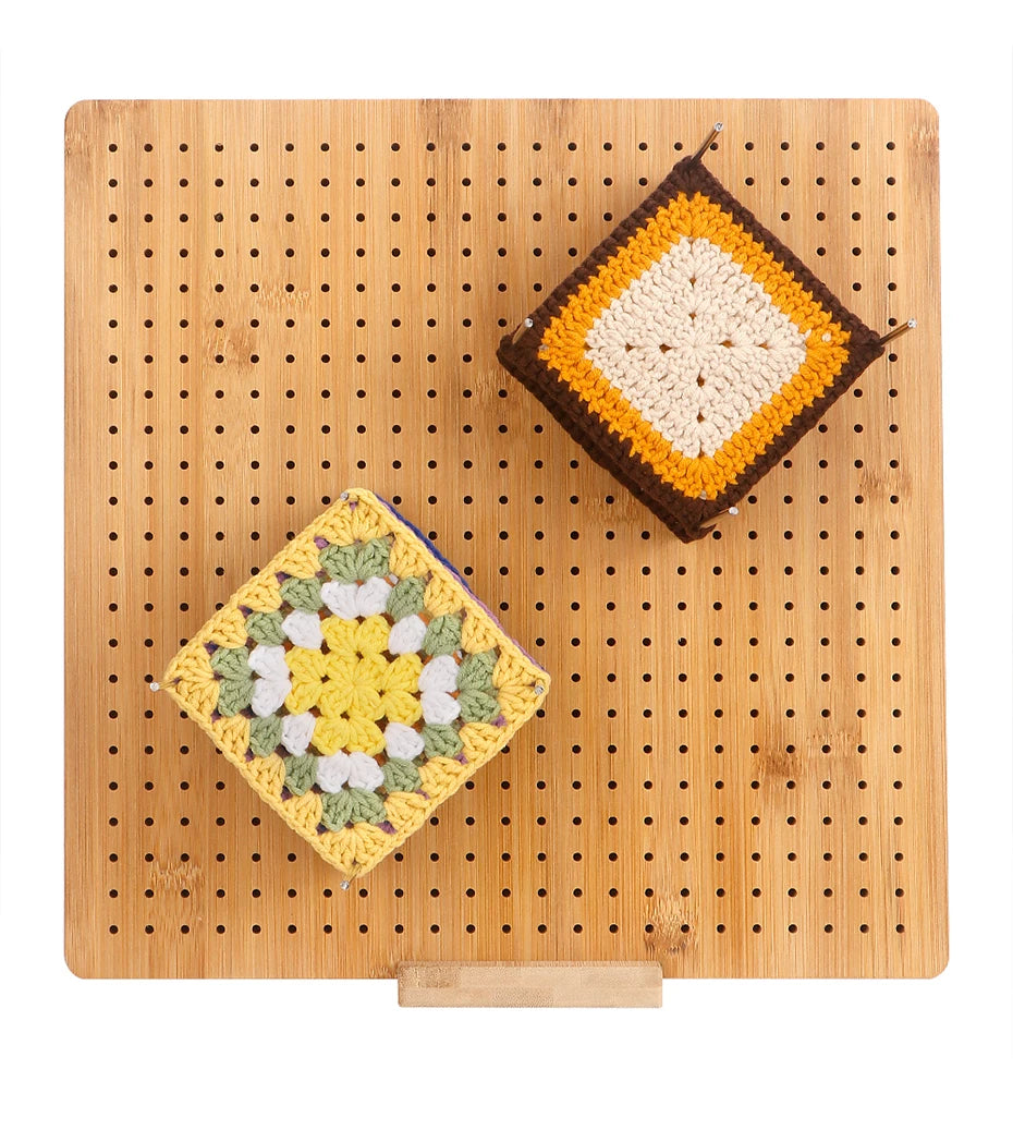 A square crochet piece featuring yellow, white, and green yarn is displayed on a wooden board with pins and a stand.