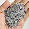 A hand holds a pile of natural tumbled stone chips in various shades of purple and green quartz for jewelry making