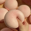 A hand is squeezing a pink, round mochi-shaped squishy stress relief toy against a light wooden surface.