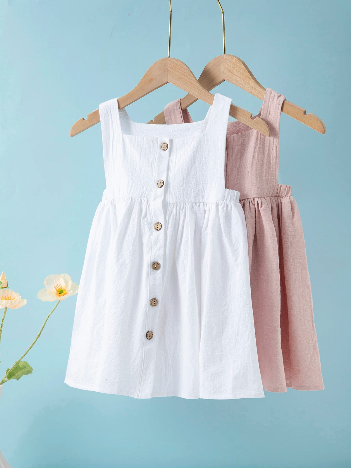 White linen jumper dress for a toddler girl hanging on a wooden hanger against a light blue background.