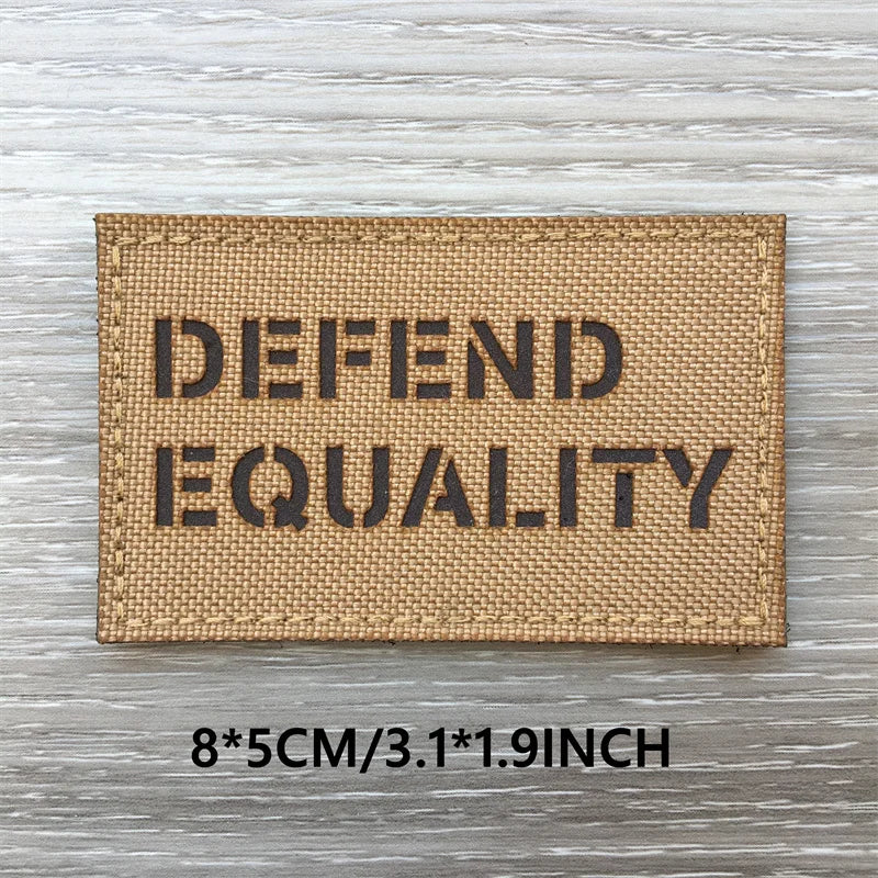 Un patch en tissu beige rectangulaire avec du texte noir indiquant DEFEND EQUALITY, affiché sur une surface en bois vieillie.