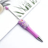 A close-up of a purple ballpoint pen with a sparkly pink sequin bead and a silver tip resting on white paper.