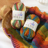 Three skeins of gradient acrylic yarn in various colors are displayed in a woven basket and on a knitted fabric.