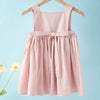 A pink linen jumper dress for a toddler girl hanging on a wooden hanger against a light blue background.