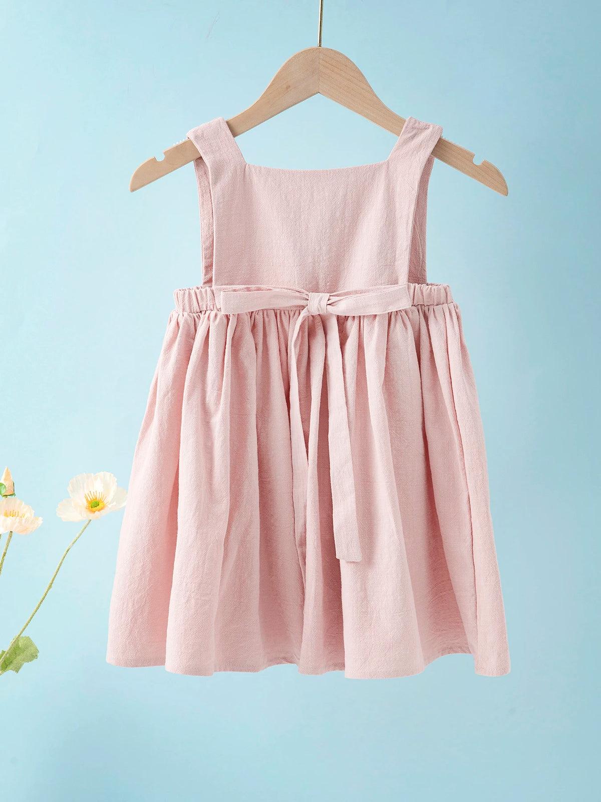 A pink linen jumper dress for a toddler girl hanging on a wooden hanger against a light blue background.