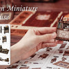 Hands assemble a miniature book nook kit featuring detailed furniture and decorative elements on a wooden surface.