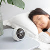 A white emoji alarm clock with a dark lens is resting on a white surface next to a person sleeping in bed.