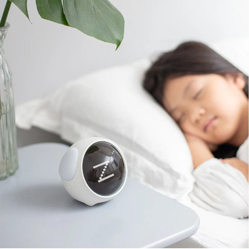 A white emoji alarm clock with a dark lens is resting on a white surface next to a person sleeping in bed.