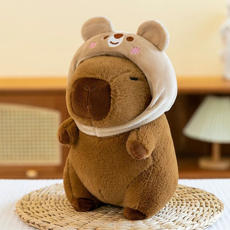 A brown capybara plush toy with large ears and a light beige hood sitting on a woven mat.