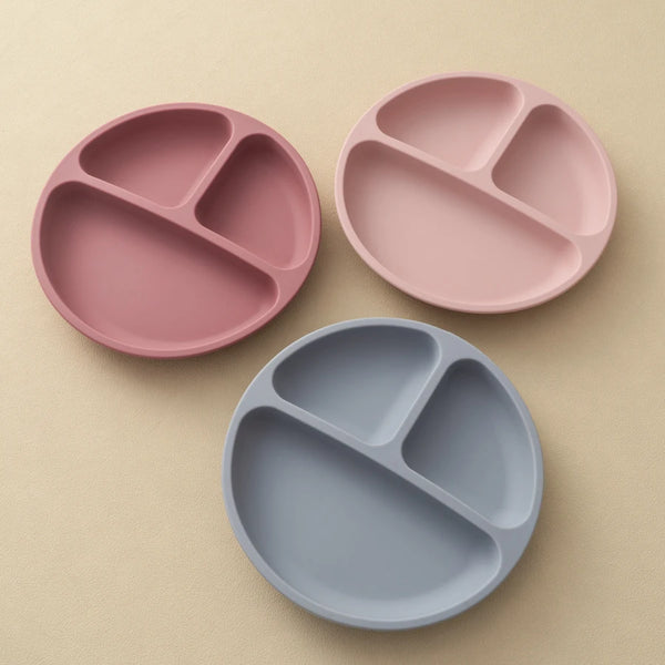 Three silicone baby sucker dining plates in pink and gray are arranged on a light beige surface.