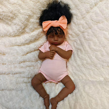 A lifelike 19-inch reborn baby doll with hand-rooted hair wearing a pink onesie, lying on a white fluffy surface.
