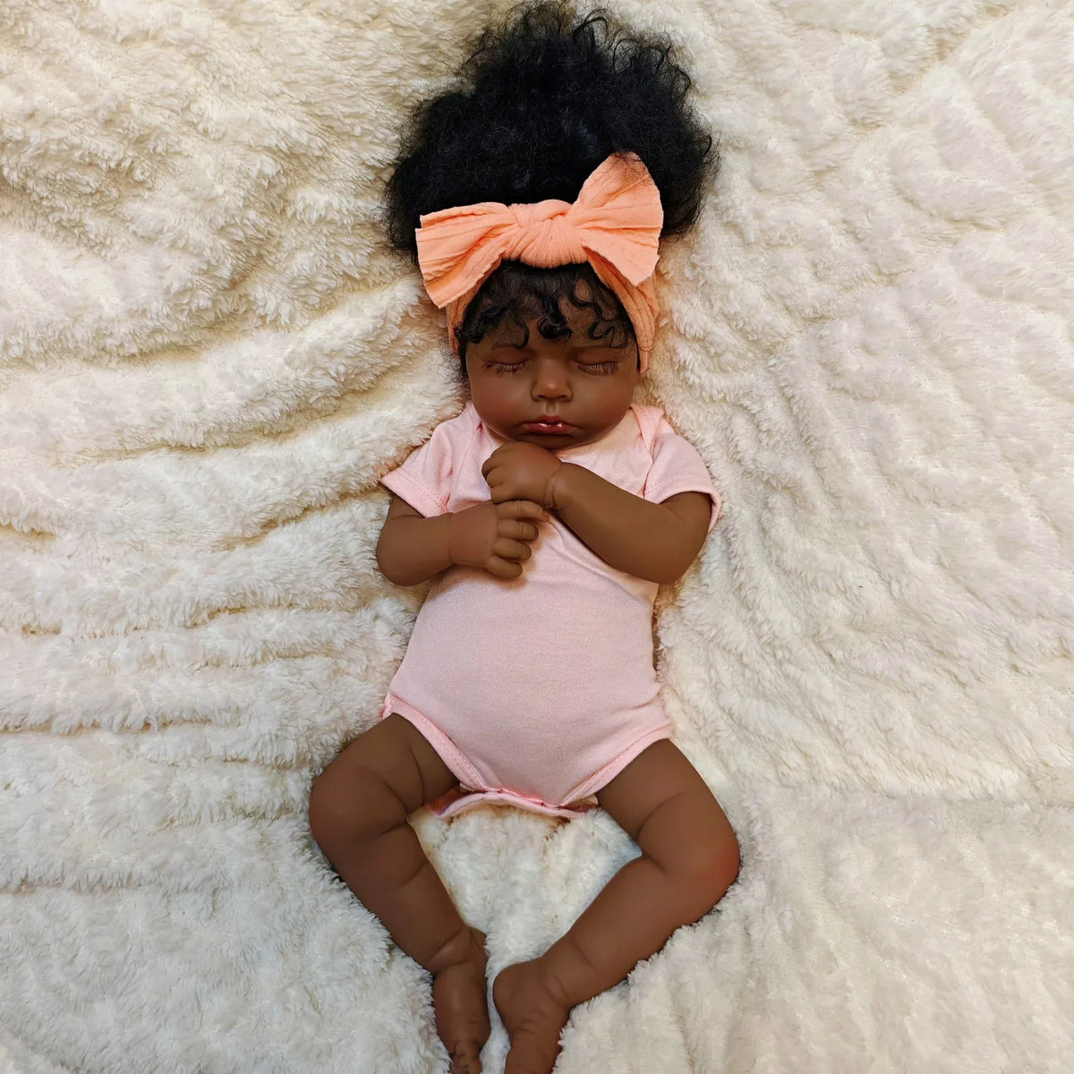 A lifelike 19-inch reborn baby doll with hand-rooted hair wearing a pink onesie, lying on a white fluffy surface.