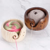 A handcrafted wooden yarn bowl with a swirl cutout holds gray and pink yarn on a white marble surface.