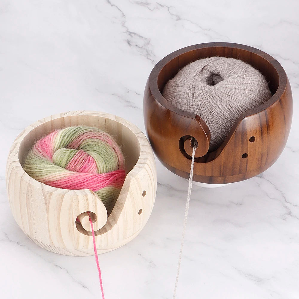 A handcrafted wooden yarn bowl with a swirl cutout holds gray and pink yarn on a white marble surface.