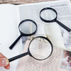 A black magnifying glass is held over text on a page, with a white candle and natural elements visible.