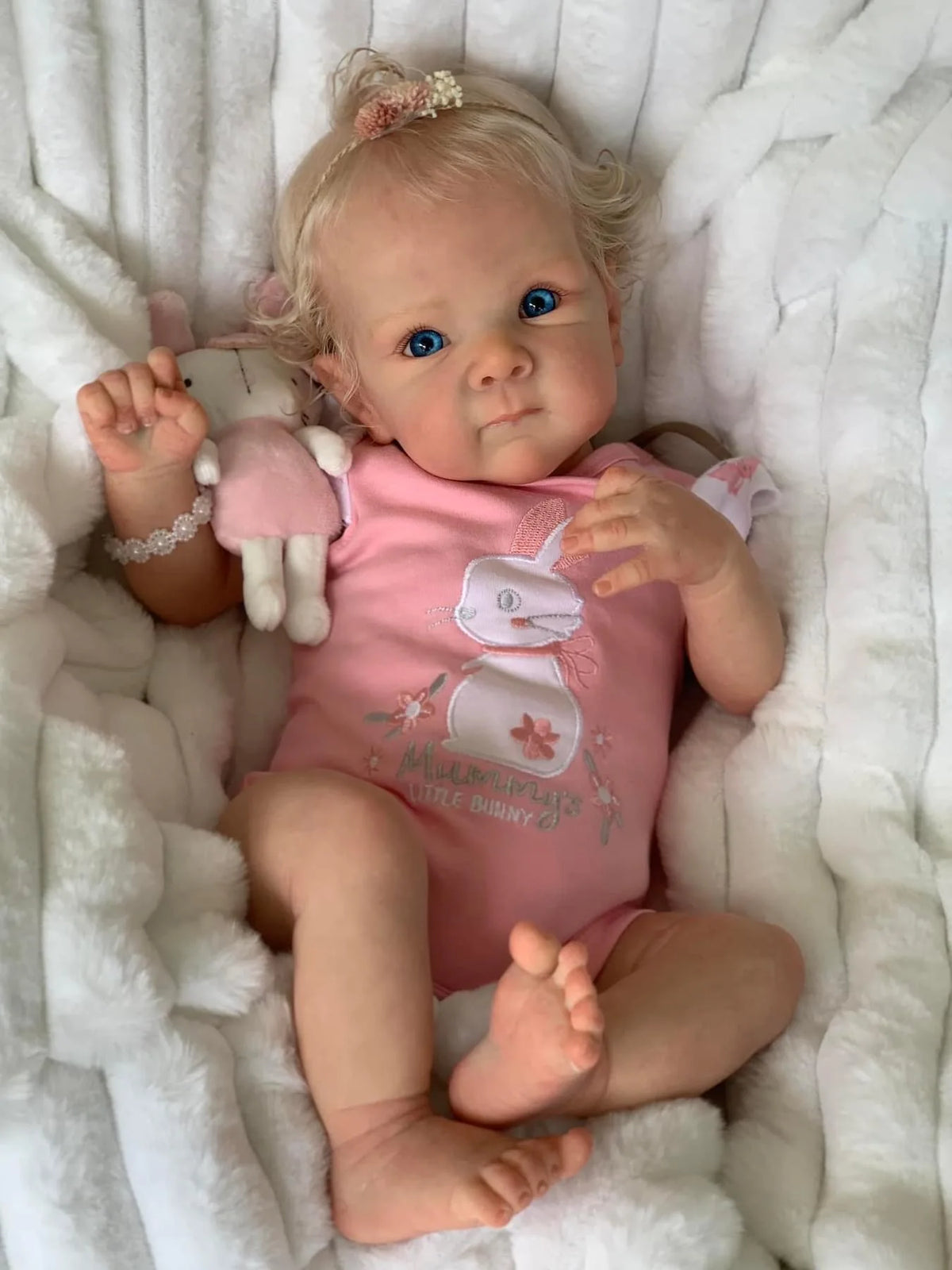 A newborn baby doll with light blonde hair is lying in a white fluffy blanket, holding a pink stuffed animal.