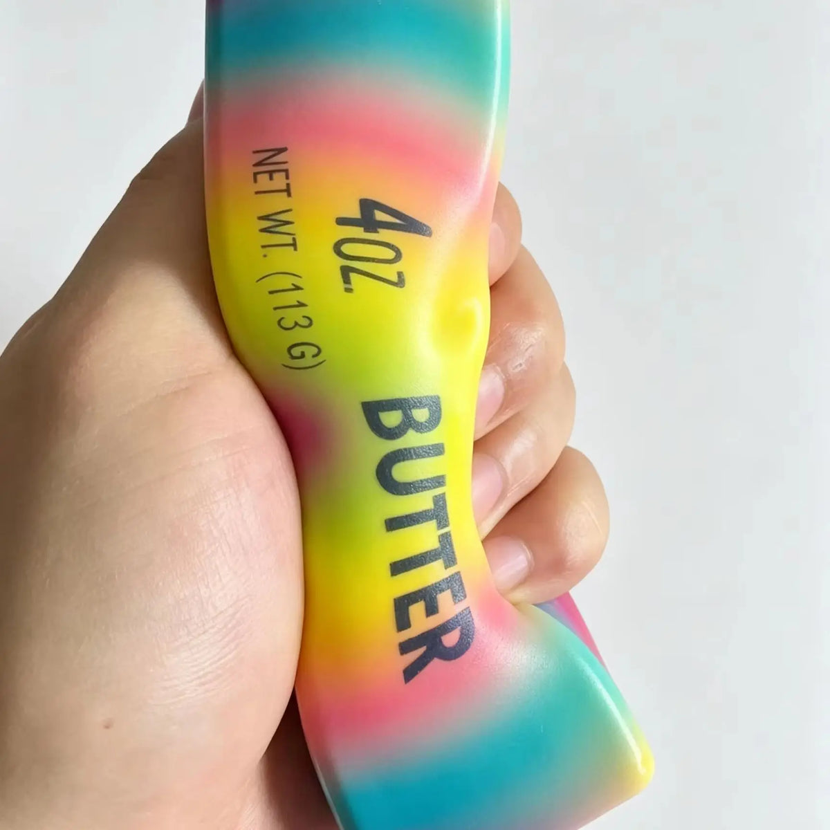 A hand holds a yellow squishy fidget toy with rainbow colored gradients and the text 40Z BUTTER NET WT 113G on it.