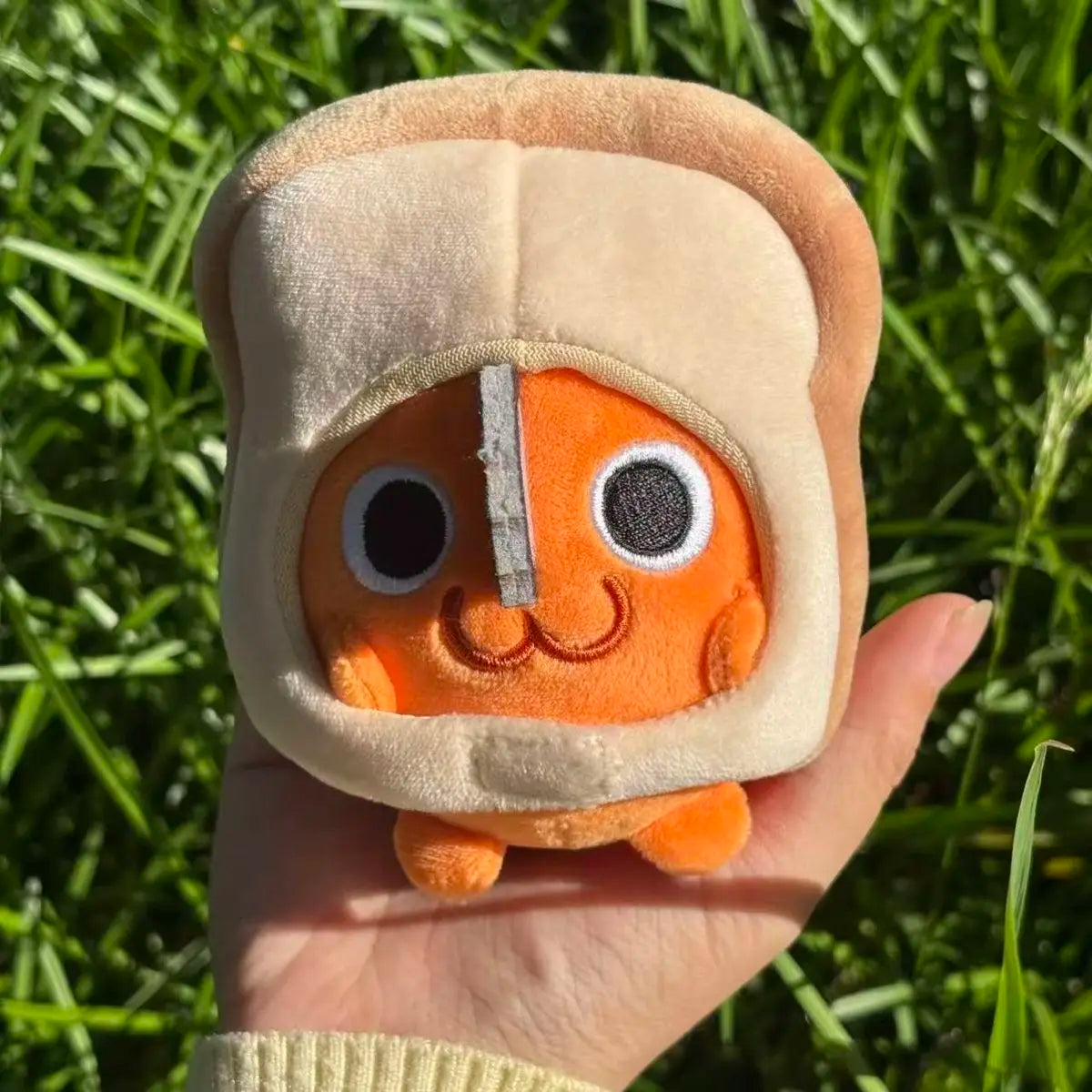 An orange Pochita plush doll with a light beige head covering is held in a hand against a green grassy background.
