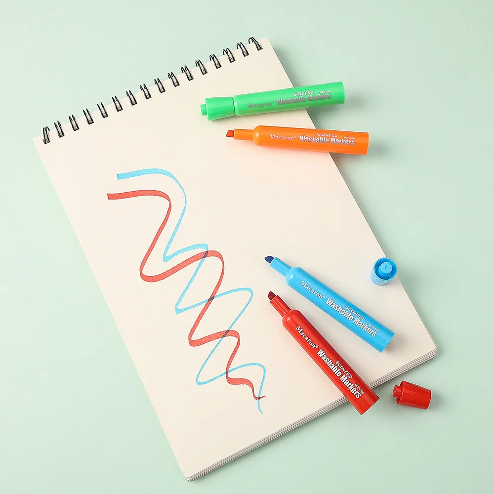 Un set de marqueurs lavables parfumés aux fruits en vert, orange et bleu sont présentés sur un carnet à spirale.
