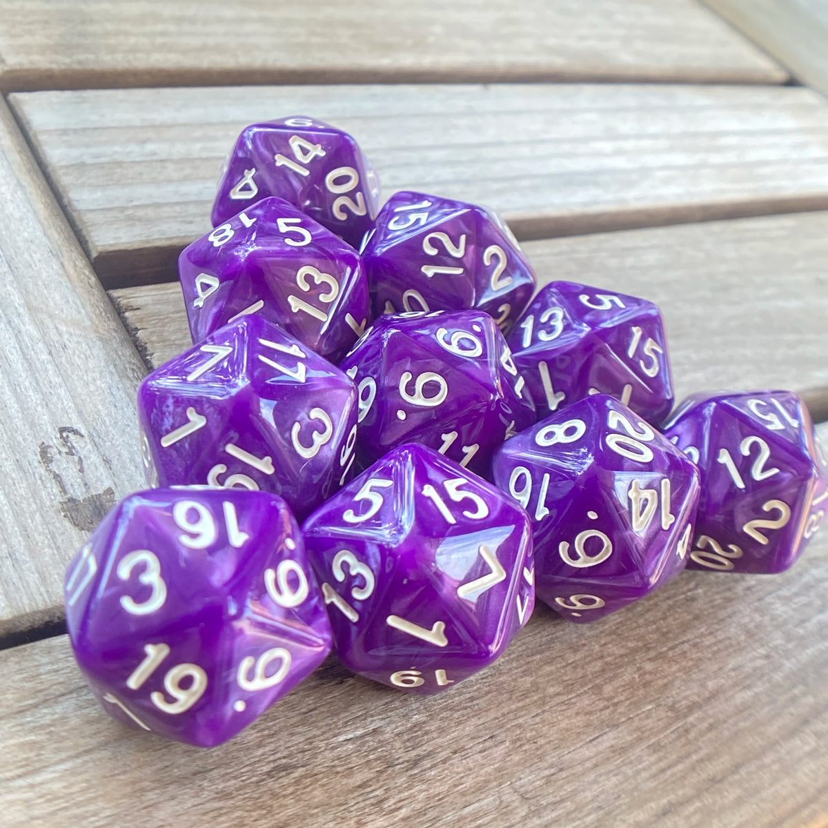 A pile of ten purple pearl acrylic D20 dice resting on a wooden surface.
