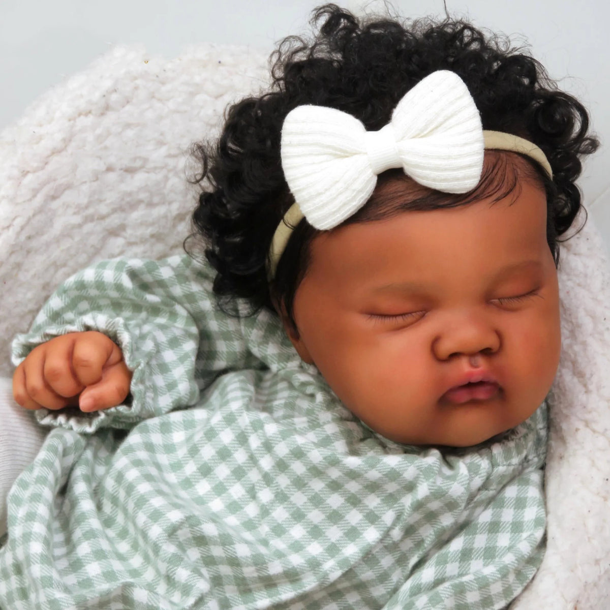 A newborn baby doll with dark curly hair wearing a white bow, wrapped in a soft white blanket.