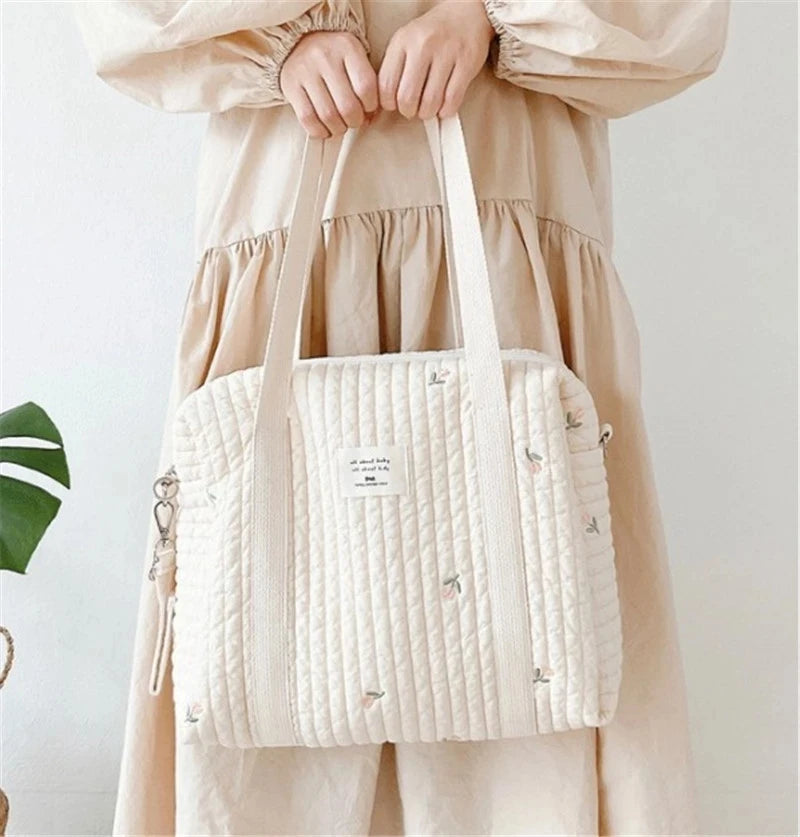 A light beige quilted diaper bag with embroidery carried by a person wearing a flowing cream-colored maternity dress.