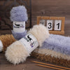 A collection of fluffy faux fur yarn in shades of purple, beige, and brown displayed on a wooden background.
