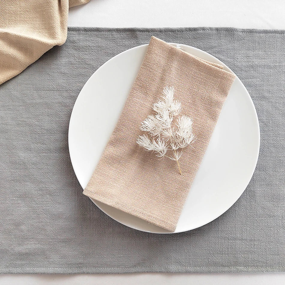 A beige cotton-linen dinner napkin folded diagonally on a white plate set against a gray linen cloth.