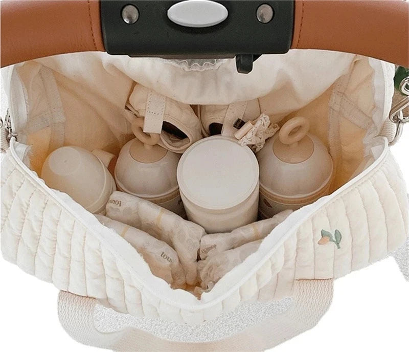 A quilted white diaper bag with embroidery is shown open, revealing soft fabric lining and various small items.