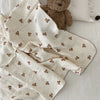 A cream muslin swaddle blanket with a subtle floral print is draped next to a brown teddy bear.