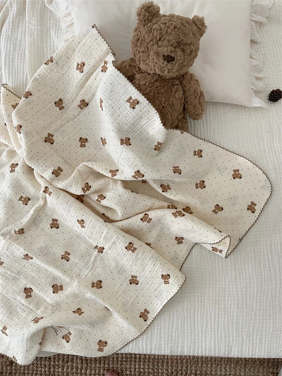 A cream muslin swaddle blanket with a subtle floral print is draped next to a brown teddy bear.