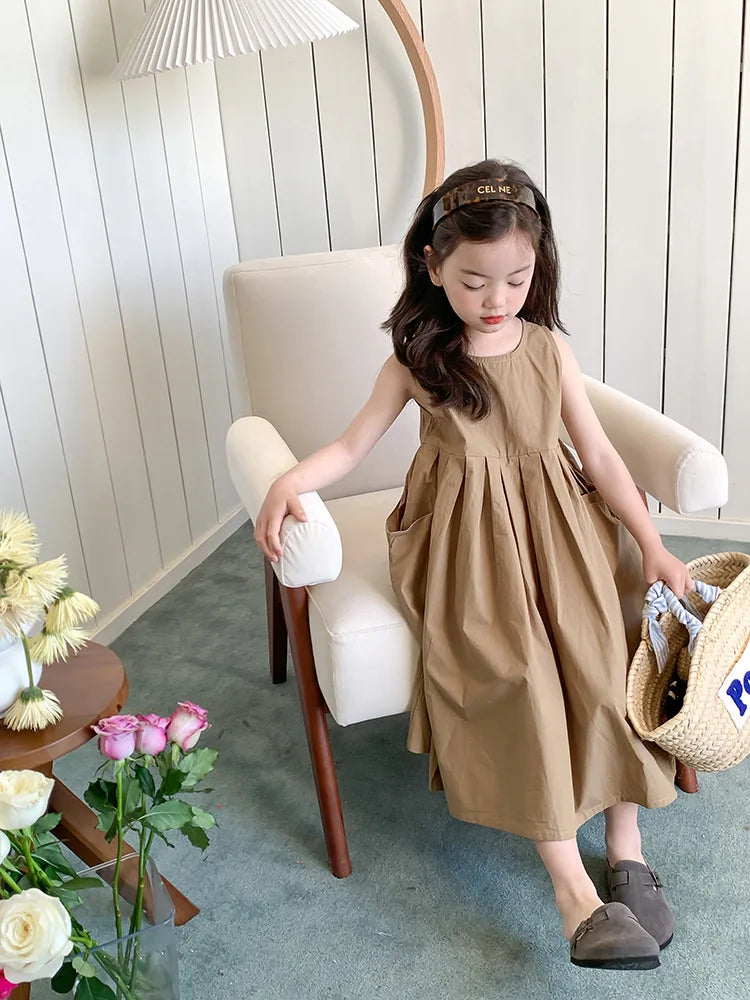 A young girl wearing a beige sleeveless cotton sundress sits in a light-colored chair.