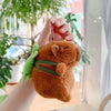 A fluffy brown capybara plush toy with green accents is being held against a blurred indoor background.