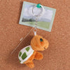 An adorable orange turtle plush toy attached to a silver keyring with a green charm, resting on brown cork board.