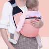A baby is being carried in an adjustable cotton ring sling baby carrier with a pink shirt and plaid skirt.