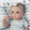 A close-up photo of a realistic reborn baby doll with painted hair wearing a gray shirt and a white bow on a striped blanket.