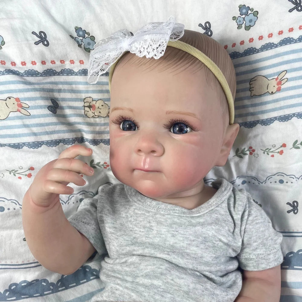 A close-up photo of a realistic reborn baby doll with painted hair wearing a gray shirt and a white bow on a striped blanket.