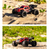 A red and black off-road RC car with large tires is photographed on sand next to green plants.